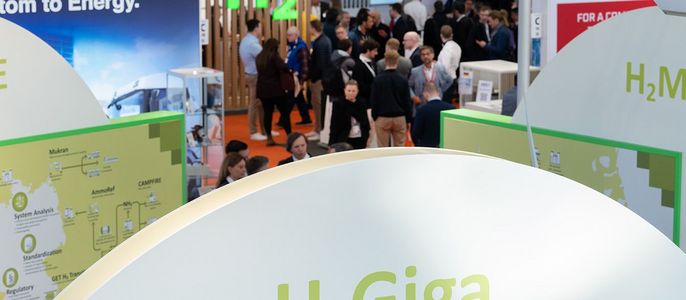 Ein Messestand, der für "H2 Giga", eine wasserstoffbasierte Energielösung, wirbt. Der Stand ist in Grün und Weiß gehalten, mit einer Deutschlandkarte, auf der die Wasserstoffproduktion und der Wasserstoffverbrauch dargestellt sind. Um ihn herum sind mehrere Personen versammelt, und im Hintergrund sind weitere Stände und Banner zu den Themen Energie und Nachhaltigkeit zu sehen.