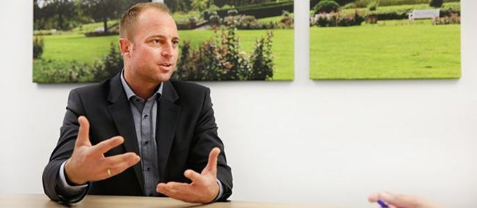 Ein Mann im Anzug, der mit seinen Händen gestikuliert, in einem Raum mit Bildern eines Gartens an der Wand.