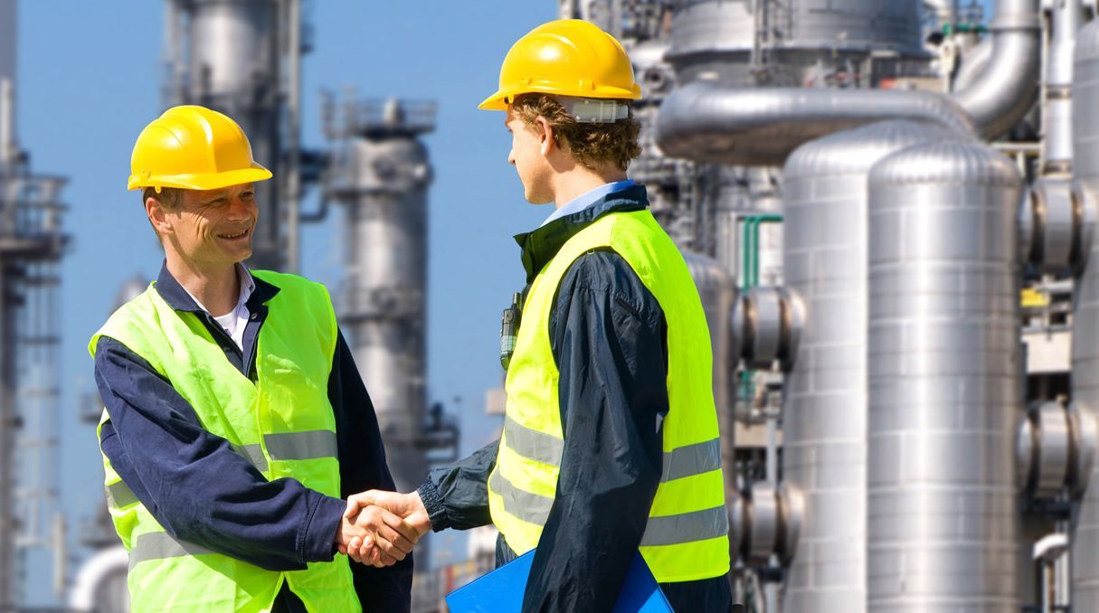 Zwei Männer in Schutzkleidung geben sich vor einer großen Industrieanlage die Hand.