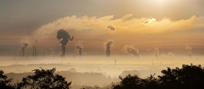 Eine weite Landschaft mit zahlreichen Schornsteinen, die vor dem Hintergrund der untergehenden Sonne Rauch ausstoßen und eine dunstige Atmosphäre am Horizont erzeugen.