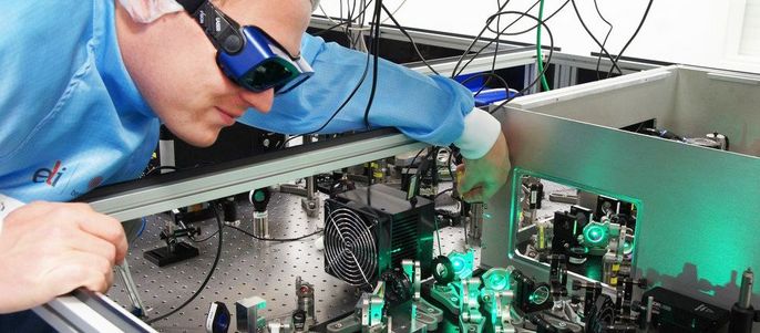 Eine Person in einem blauen Laborkittel, die eine Schutzbrille trägt und einen komplexen Apparat mit grün leuchtenden Bauteilen genau untersucht.