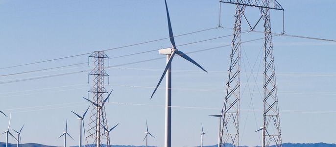 Ein riesiges offenes Feld mit mehreren Windkraftwerken, die durch Stromleitungen miteinander verbunden sind, vor einer Kulisse aus sanften Hügeln und einem klaren blauen Himmel.