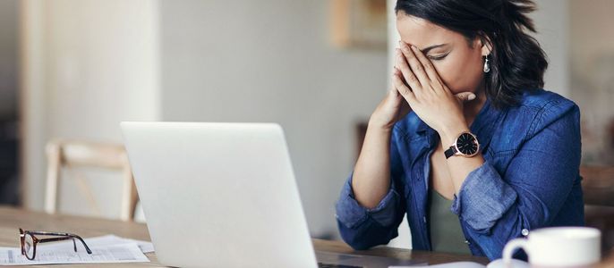 Eine Frau mit dunklem Haar und blauem Hemd sitzt an einem Tisch mit einem Laptop, einer Brille und einer Tasse. Ihre Hände bedecken ihr Gesicht in einer Geste der Verzweiflung.