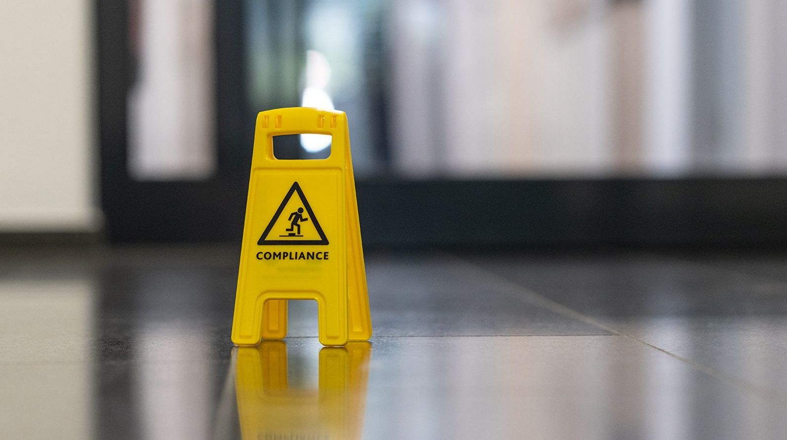 Ein gelbes "COMPLIANCE"-Schild auf einem glänzenden Boden, mit einem unscharfen Hintergrund eines Gebäudes.