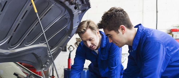 Zwei Männer in blauen Uniformen arbeiten in einer Werkstatt an einem Automotor.