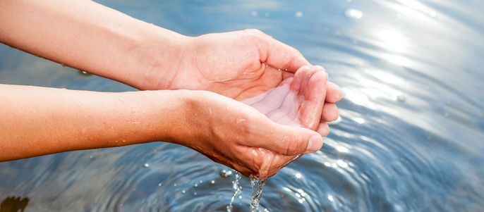Die Hände einer Person, die klares Wasser halten, das im Wasser darunter Wellen verursacht.