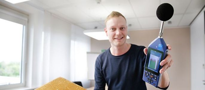 Ein Mann hält ein Gerät mit einem Mikrofon in der Hand, daneben ein Holzobjekt mit einem runden Loch.